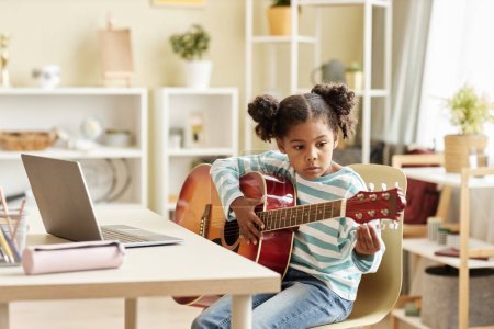 Photo for Portrait of black little girl playing guitar at home and watching online music lesson in sunny room , copy space - Royalty Free Image