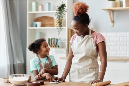 Foto de Retrato de la cintura hacia arriba de la madre negra feliz y la hija que cocinan juntas en la cocina acogedora y usando delantales - Imagen libre de derechos
