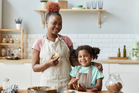 Photo for Waist up portrait of happy mother and daughter tasting fresh homemade cookies and looking at camera while enjoying baking together in kitchen - Royalty Free Image