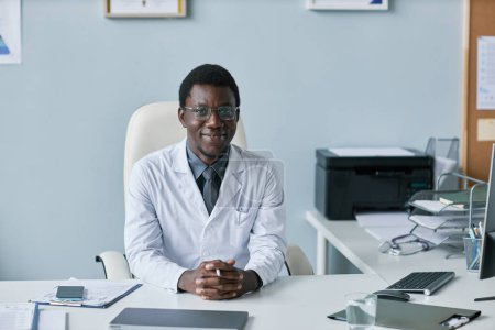 Téléchargez les photos : Minimale vue de face portrait d'un médecin noir souriant regardant la caméra alors qu'il était assis au bureau dans le bureau de la clinique, espace de copie - en image libre de droit