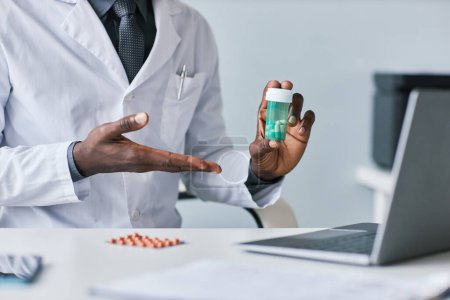 Foto de Primer plano del médico negro sosteniendo la botella de pastillas y explicando los efectos secundarios de la medicación durante la consulta en línea - Imagen libre de derechos