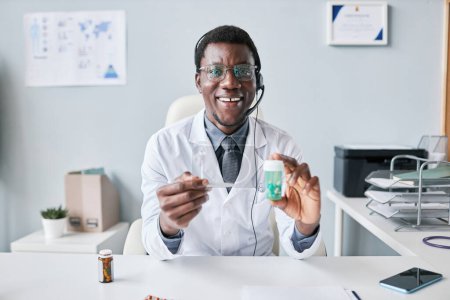 Foto de Retrato del médico negro sosteniendo una botella de pastillas y usando auriculares en el lugar de trabajo en la oficina - Imagen libre de derechos