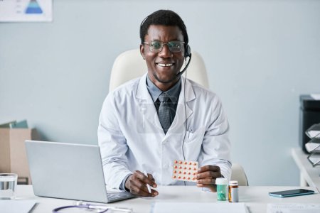 Téléchargez les photos : Portrait de médecin afro-américain souriant tenant des comprimés et portant un casque sur le lieu de travail au bureau - en image libre de droit
