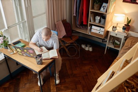 Téléchargez les photos : Portrait en grand angle d'une jeune femme chauve faisant ses devoirs dans un dortoir et prenant des notes du manuel, espace de copie - en image libre de droit