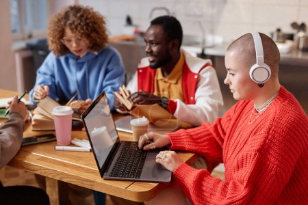 Photo for Multiethnic group of young people studying together at table in college dorm., focus on bald young woman using laptop, copy space - Royalty Free Image