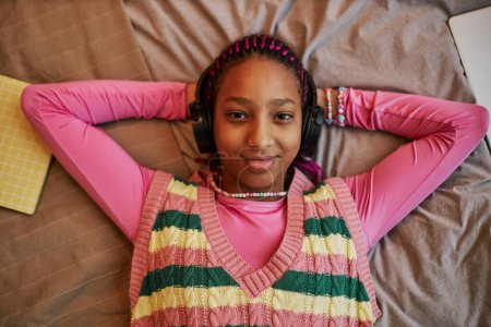 Foto de Retrato de vista superior de una adolescente negra escuchando música con auriculares mientras está acostada en la cama y vistiendo de rosa - Imagen libre de derechos