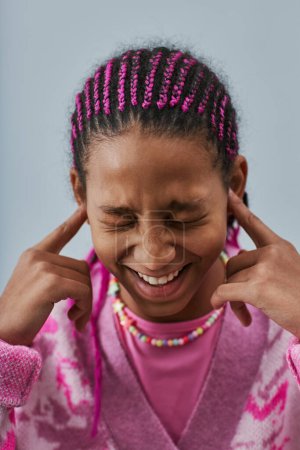 Photo for Close up portrait of expressive black teenage girl wearing pink and closing ears against blue background - Royalty Free Image