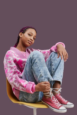 Téléchargez les photos : Portrait vertical pleine longueur d'adolescente noire portant des vêtements décontractés à la mode et posant assis sur une chaise avec les pieds levés - en image libre de droit