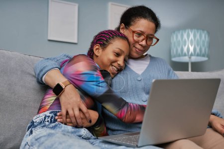 Téléchargez les photos : Portrait d'adolescente noire utilisant un ordinateur portable tout en câlinant avec maman sur le canapé - en image libre de droit
