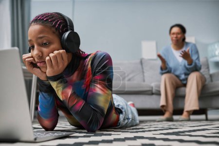 Téléchargez les photos : Portrait d'une adolescente noire utilisant un ordinateur portable et portant un casque tout en étant couchée sur le sol - en image libre de droit