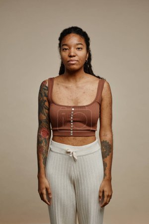 Foto de Retrato mínimo de una joven mujer negra mirando la cámara de pie sobre fondo beige neutro con enfoque en la textura de la piel real con manchas y cicatrices de acné - Imagen libre de derechos