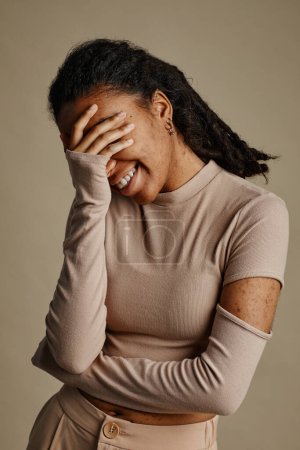 Téléchargez les photos : Portrait vertical de jeune femme noire riant sincèrement et couvrant le visage dans des tons neutres - en image libre de droit