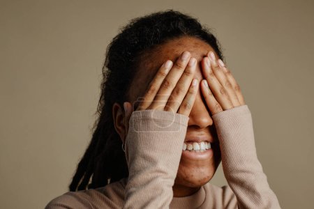 Foto de Primer plano de la joven mujer negra sonriendo felizmente y cubriendo los ojos con las manos, se centran en la textura de la piel de la cara con cicatrices de acné, espacio de copia - Imagen libre de derechos