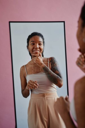 Photo for Vertical portrait of young black woman with tattoos and acne scars looking in mirror smiling confidently - Royalty Free Image