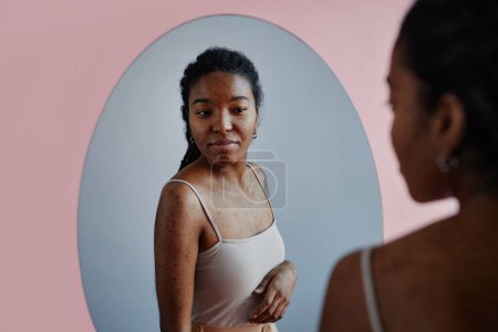 Téléchargez les photos : Portrait franc de jeune femme noire avec des cicatrices d'acné regardant dans le miroir et souriant - en image libre de droit
