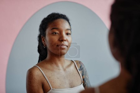 Foto de Joven mujer negra con tatuajes y cicatrices de acné mirando en el espejo sonriendo - Imagen libre de derechos