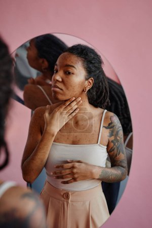 Téléchargez les photos : Portrait vertical de jeune femme noire avec des cicatrices d'acné regardant dans le miroir d'apparence précaire - en image libre de droit