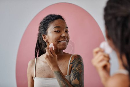 Téléchargez les photos : Candid portrait minimal de jeune femme noire avec des cicatrices d'acné à l'aide d'un masseur visage pendant la routine de beauté et souriant - en image libre de droit