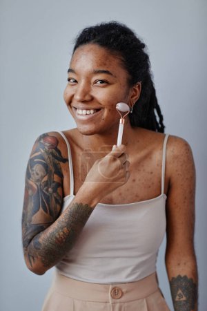 Téléchargez les photos : Portrait vertical de jeune femme noire avec des cicatrices d'acné et des tatouages à l'aide d'un masseur du visage et souriant, minimal - en image libre de droit