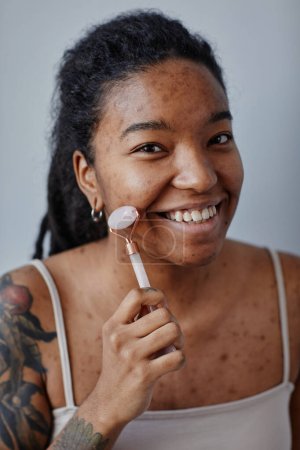 Téléchargez les photos : Portrait vertical rapproché d'une jeune femme noire utilisant un masseur facial et souriant, minimal - en image libre de droit