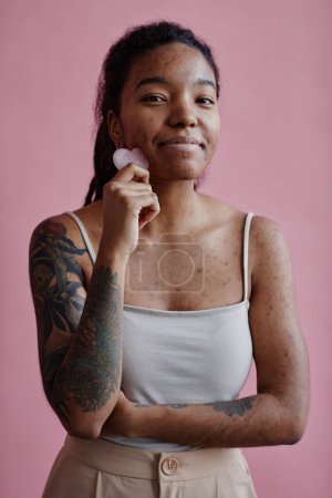 Téléchargez les photos : Portrait vertical de jeune femme noire avec des cicatrices d'acné à l'aide d'un masseur facial et souriant contre le rose - en image libre de droit