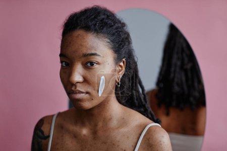 Téléchargez les photos : Portrait franc de jeune femme noire avec une tache de crème visage regardant la caméra par miroir - en image libre de droit