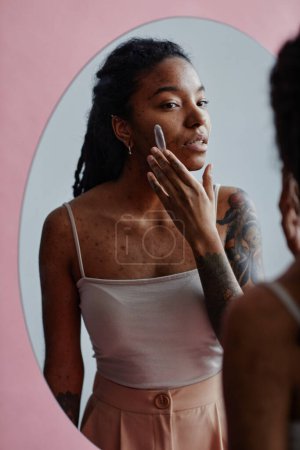 Téléchargez les photos : Portrait vertical de jeune femme noire avec des cicatrices d'acné en utilisant la crème visage regardant dans le miroir - en image libre de droit