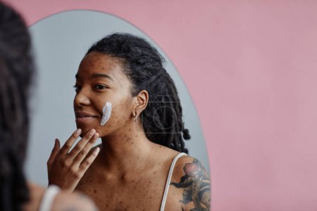 Téléchargez les photos : Portrait franc de jeune femme noire avec des cicatrices d'acné en utilisant la crème visage regardant dans la réflexion miroir, espace de copie - en image libre de droit
