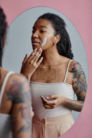 Téléchargez les photos : Portrait vertical franc de jeune femme noire avec des cicatrices d'acné en utilisant la crème visage par miroir - en image libre de droit