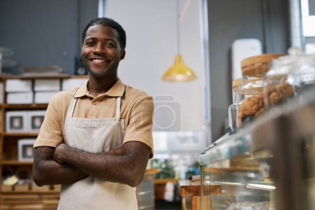 Téléchargez les photos : Joyeux petit travailleur de boulangerie confiant debout au comptoir et regardant la caméra - en image libre de droit