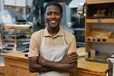 Téléchargez les photos : Portrait de boulanger heureux dans le tablier debout avec les bras croisés et souriant à la caméra - en image libre de droit