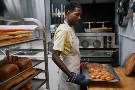 Téléchargez les photos : Travailleur de boulangerie mettant un plateau chaud avec du pain fraîchement cuit sur la table pour le laisser refroidir - en image libre de droit