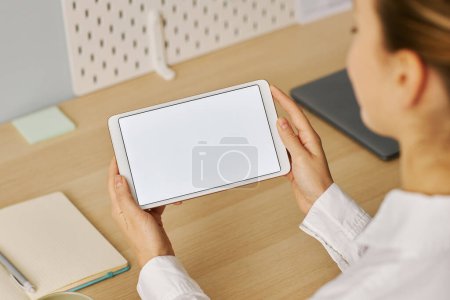 Foto de Vista de ángulo alto de la mujer joven sosteniendo tableta con pantalla blanca maqueta en el lugar de trabajo - Imagen libre de derechos