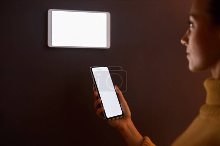 Téléchargez les photos : Femme utilisant le système numérique de maison intelligente avec des maquettes d'écran sur le mur et le téléphone - en image libre de droit