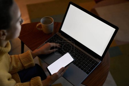 Téléchargez les photos : Gros plan d'angle élevé de la jeune femme utilisant des dispositifs avec des maquettes d'écran blanc dans le cadre sombre de la maison - en image libre de droit