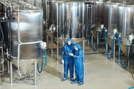 Téléchargez les photos : Vue grand angle à l'atelier d'usine avec deux travailleurs en équipement de protection par des réservoirs de stockage, espace de copie - en image libre de droit