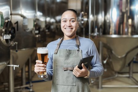 Téléchargez les photos : Portrait taille haute d'une femme souriante tenant un verre de bière et regardant la caméra tout en travaillant à l'usine de brassage - en image libre de droit