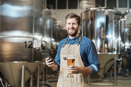 Téléchargez les photos : Portrait taille haute de brasseur barbu tenant verre de bière et regardant la caméra tout en se tenant à l'usine de brassage - en image libre de droit