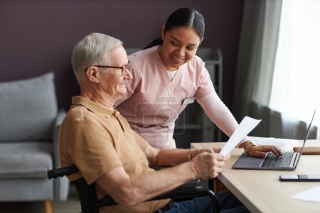 Photo for Senior man with disability learning to pay bills online with laptop together with social worker - Royalty Free Image