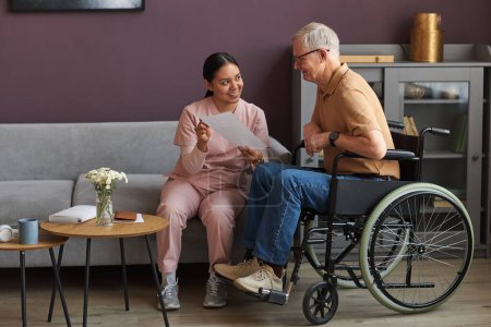 Photo for Young caregiver explaining the way of payment of bills to senior man with disability while they sitting in the room - Royalty Free Image