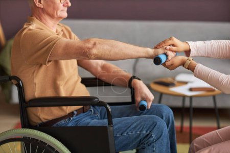 Foto de Hombre mayor con discapacidad sentado en silla de ruedas y haciendo ejercicio con pesas con asistente social ayudándole - Imagen libre de derechos