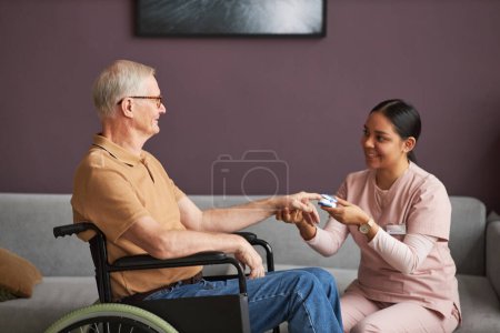 Photo for Young social worker checking pressure of senior man with disability with special medical equipment during her visit to home - Royalty Free Image