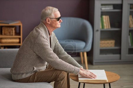 Téléchargez les photos : Homme âgé aux lunettes sombres avec cécité lisant un livre à table dans le salon - en image libre de droit