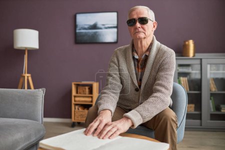 Foto de Hombre mayor ciego en anteojos leyendo un libro en Braille en la mesa de la sala de estar - Imagen libre de derechos