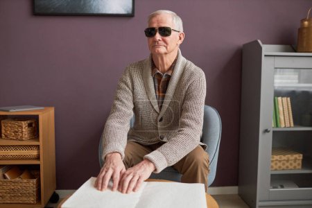 Téléchargez les photos : Aveugle âgé aux lunettes sombres apprenant à lire un livre en braille à la maison - en image libre de droit