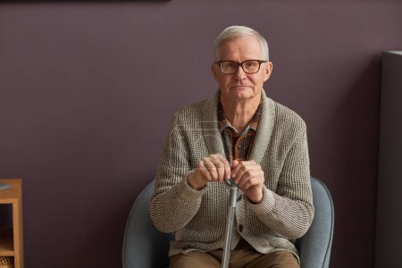 Téléchargez les photos : Portrait d'un homme âgé en lunettes avec un bâton assis sur un fauteuil et regardant la caméra - en image libre de droit
