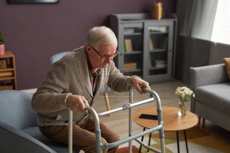 Téléchargez les photos : Homme âgé utilisant marcheur pour se lever du canapé, il passe du temps dans le salon à la maison - en image libre de droit