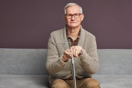 Foto de Retrato de anciano en gafas con palo sentado en el sofá y mirando a la cámara - Imagen libre de derechos