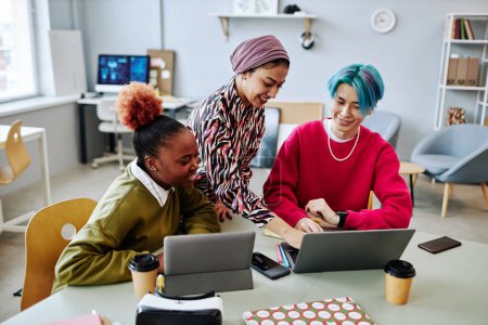 Téléchargez les photos : Groupe diversifié de jeunes créatifs utilisant un ordinateur portable lors d'une réunion au bureau et souriant joyeusement - en image libre de droit