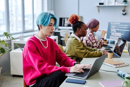 Téléchargez les photos : Une équipe diversifiée de développeurs de logiciels de génération Z travaillant dans le bureau se concentre sur le jeune homme asiatique aux cheveux colorés en utilisant l'ordinateur au premier plan - en image libre de droit